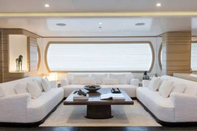 Three white sofas and a dark wooden coffee table sit in the foreground of a modern, neutral-toned living room with a large window in the background.