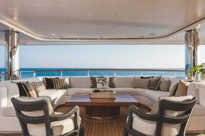 A luxurious white sectional sofa and two armchairs surround a wooden coffee table on a yacht deck, overlooking a calm blue ocean under a clear sky.