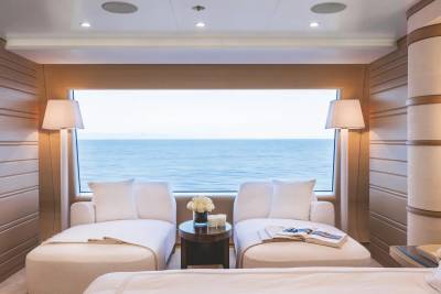 Two white lounge chairs and a small side table sit in a modern room facing a large window with a view of the calm blue ocean.