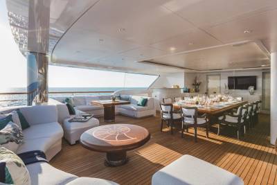 A luxurious yacht deck features a white lounge area and a round wooden table in the foreground, with a formal dining set and a view of the ocean in the background.