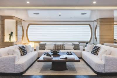 A modern living area features white sofas and a dark wooden coffee table in the foreground, set against a background of neutral-toned walls and a large window with white blinds.