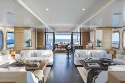 A luxurious yacht interior features white sofas and wooden coffee tables in the foreground, leading toward an open deck with a view of the blue ocean in the background.