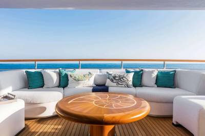 A round wooden table and a white curved sofa with teal pillows sit on a wooden deck overlooking a calm blue ocean under a clear sky.