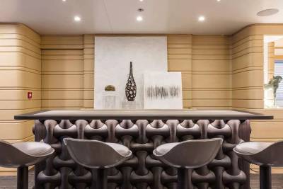 Four modern grey bar stools sit before a uniquely textured dark brown counter, set against a backdrop of ribbed beige walls and minimalist art.