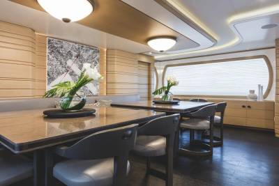 Two modern dining tables with grey chairs and floral centerpieces sit in the foreground of a luxurious room featuring beige paneled walls and a large window in the background.