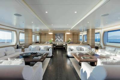White modern sofas and wooden coffee tables fill the foreground of a luxurious yacht interior, with large windows overlooking the ocean in the background.