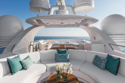 A white curved outdoor sofa with teal pillows and a flower arrangement sits in the foreground of a luxury yacht deck overlooking a calm blue ocean.