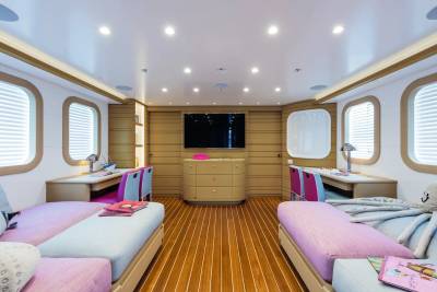 Two twin beds with purple and blue bedding sit in the foreground of a modern yacht cabin featuring wooden floors, desks, and a television on the back wall.