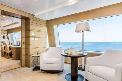 Two white armchairs and a small round table sit in a luxurious room, with a large window overlooking a calm blue ocean in the background.