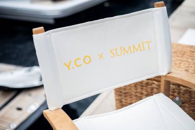 A white director's chair with yellow "Y.CO x SUMMIT" embroidery sits in the foreground against a blurred background of a woven basket and dark flooring.