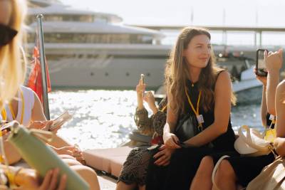 A woman in a black dress sits on a deck in the foreground, while a large white yacht and shimmering water fill the background.