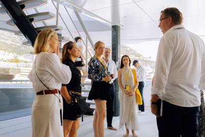 A group of people in professional attire stand and converse in a modern, bright interior with a staircase and a scenic coastal view in the background.