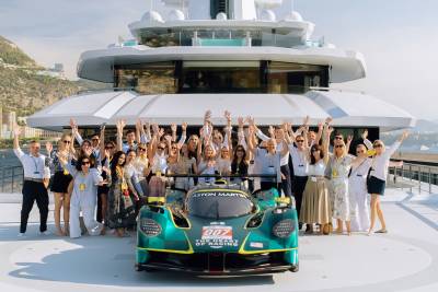 A group of people pose with a green Aston Martin race car on the deck of a massive white luxury yacht.