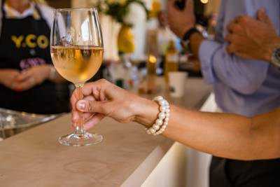 A hand wearing a pearl bracelet holds a glass of white wine over a counter, with a person in a black apron and another in a blue shirt blurred in the background.