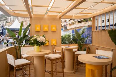 Small round tables and white chairs sit in a light-wood lounge area, with green plants and a city skyline visible in the background.