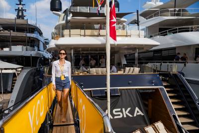 A woman in a white shirt and denim shorts walks down a gangway in the foreground, with several luxury yachts docked in the background.