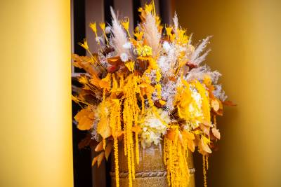 A vibrant bouquet of yellow and orange dried flowers sits in a golden vase against a bright yellow wall.
