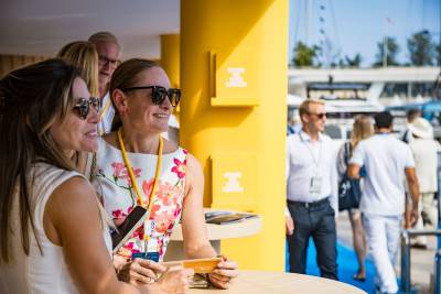 Three women smile and converse in the foreground beside a bright yellow pillar, with several people and boats visible in the sunny outdoor background.