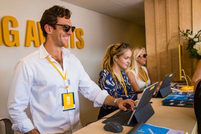 Three people wearing yellow lanyards work on tablets at a desk in front of a wall with large yellow lettering.