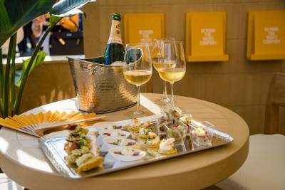 A variety of gourmet appetizers and glasses of white wine sit on a round table next to a champagne bucket, with yellow branded signs on the wall in the background.
