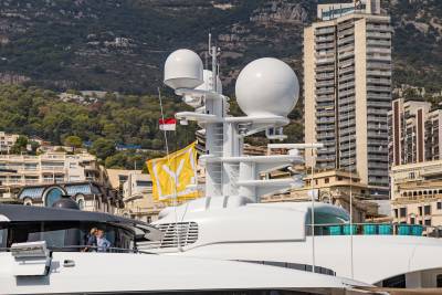A white luxury yacht with large radar domes stands in the foreground against a backdrop of tall buildings and a steep, forested mountain.