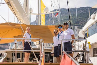 Four crew members in white shirts and dark shorts stand on a yacht deck against a backdrop of a mountain and city skyline.