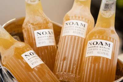 Four chilled bottles of Noam beer sit in a metal ice bucket against a blurred, neutral background.