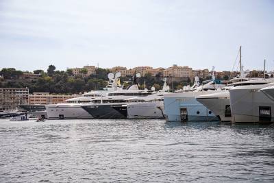 Several luxury yachts are docked in a harbor with a hillside town and buildings in the background.