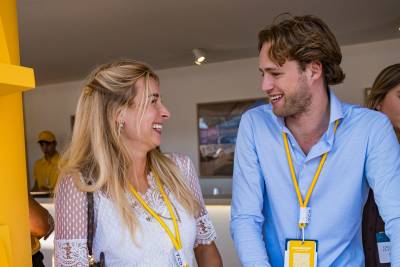 A smiling blonde woman and a man in a blue shirt talk in the foreground against a blurred indoor background.