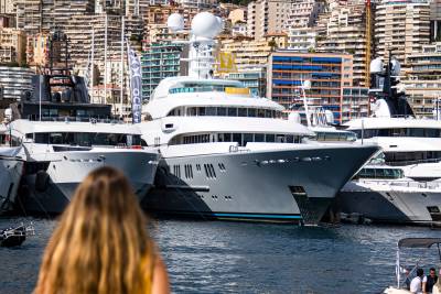 A woman with long blonde hair looks toward several massive luxury yachts docked in a harbor against a backdrop of dense city buildings.