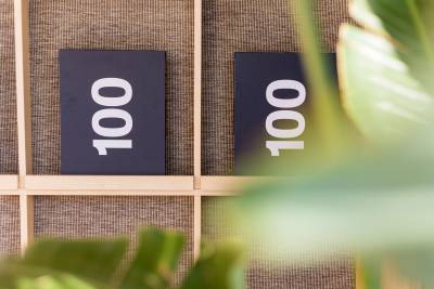 Blurry green leaves frame the foreground of two black signs with the number "100" set against a textured brown wall with light wood framing.