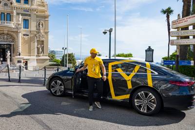 A man in a yellow shirt and cap steps out of a black car with yellow graphics, parked in front of an ornate classical building.