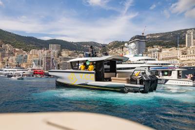 A modern white and black boat with "Y.CO" branding cruises through blue water, with a backdrop of luxury yachts and a densely populated hillside city.