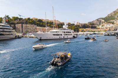 Various boats and yachts navigate a blue harbor with a cityscape and mountains in the background.