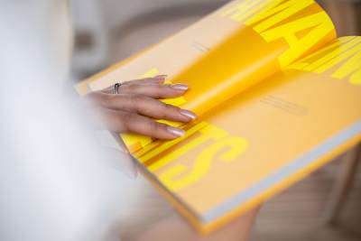 A hand with a ring and polished nails rests on a bright yellow open magazine against a blurred, neutral background.