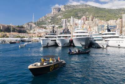 A small black motorboat with passengers floats in the foreground of a harbor filled with luxury yachts, set against a backdrop of a coastal city and steep mountains.