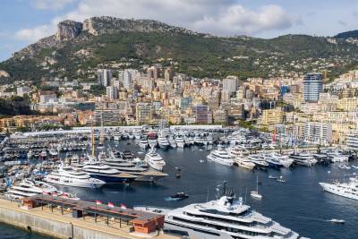 Numerous luxury yachts fill a bustling harbor in the foreground, set against a backdrop of a densely populated hillside city and towering green mountains.