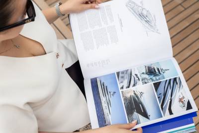 A woman in a white top looks through a brochure of luxury yachts while standing on a wooden deck.
