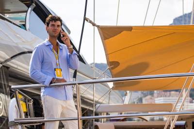 A man in a light blue shirt and white trousers talks on a phone while leaning on a yacht railing, with a large yellow sunshade and a coastal landscape in the background.