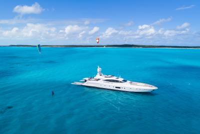 A sleek white yacht floats in turquoise waters with a kite surfer nearby, set against a distant coastline and a bright blue sky.