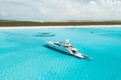 A luxury white yacht floats in crystal clear turquoise water with a sandy beach and green shrubbery in the background.