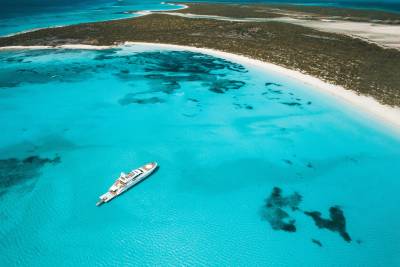 A white yacht floats in crystal clear turquoise water near a white sandy beach and a scrub-covered island.