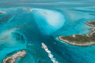 A white yacht cruises through turquoise waters in the foreground, surrounded by rocky islands and shallow coral reefs in the background.