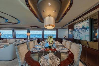 An elegant dining table set for a meal occupies the foreground of a luxury yacht interior, with a plush lounge area and ocean views visible in the background.