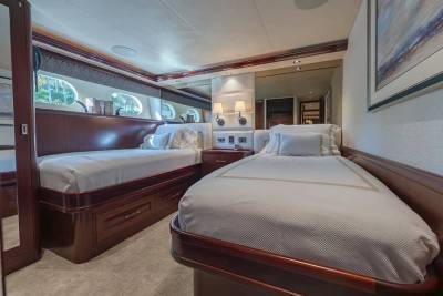 Two twin beds with white linens sit in a luxurious cabin featuring polished wood paneling and small porthole windows in the background.
