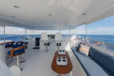 A luxurious yacht deck features a lounge area with a blue sofa and bar stools in the foreground, overlooking a calm blue ocean and a distant city skyline in the background.