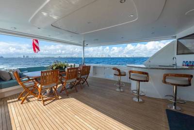 A luxury yacht deck features a dining area and bar in the foreground, overlooking a blue ocean and a distant city skyline in the background.