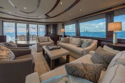 A luxurious yacht lounge with plush beige seating and a coffee table in the foreground opens up to a scenic ocean view and a distant city skyline in the background.