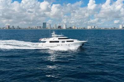 A white luxury yacht cruises through deep blue water with a city skyline and cloudy sky in the background.