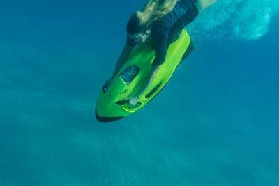 A woman dives underwater on a bright green seabob in the foreground, with another person on a similar device gliding through the blue water in the background.